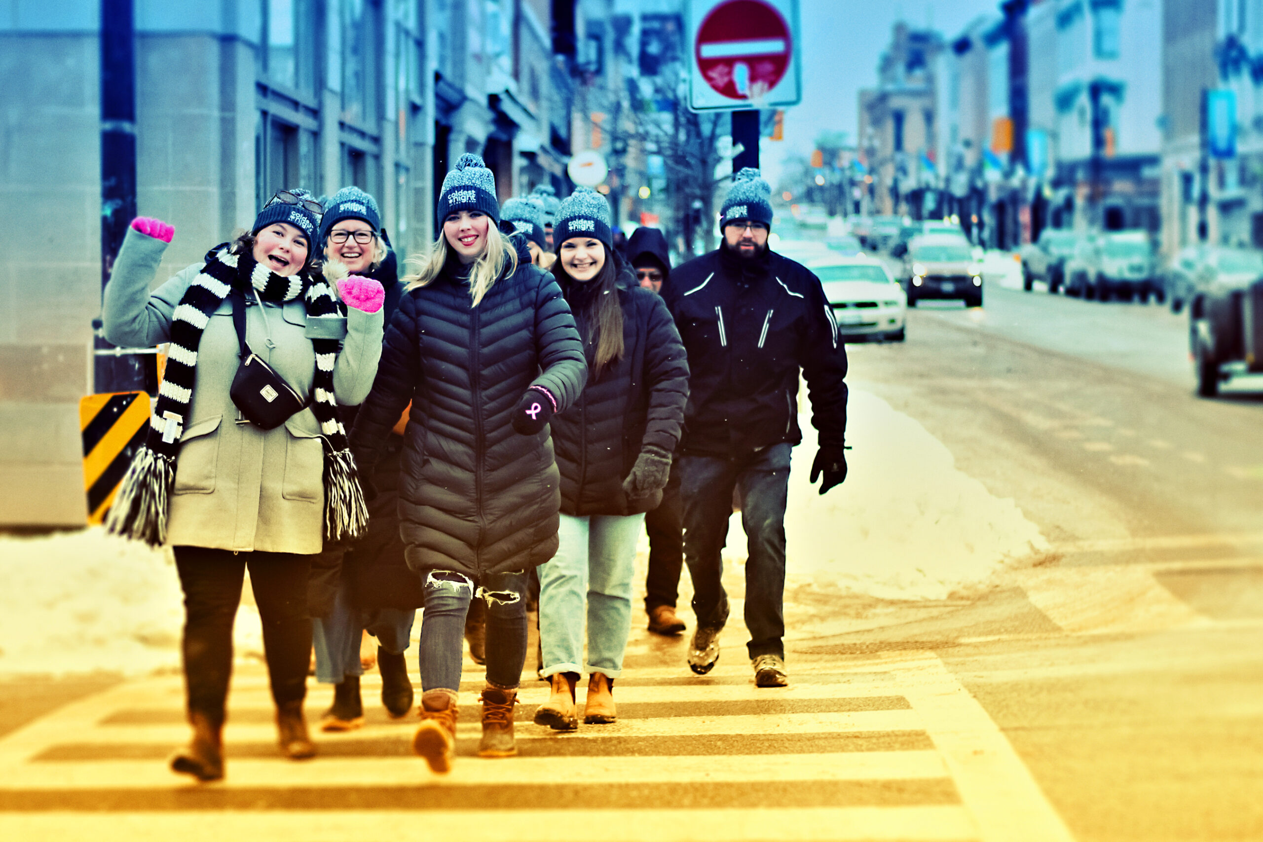 A photo of a group of people walking, crossing the street at a crosswalk. They are happy and waving.